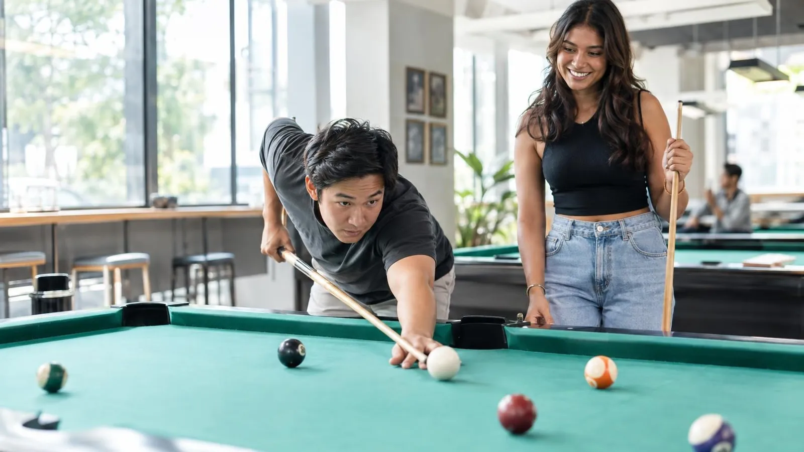 Two friends playing pool at a green-felt table in a bright modern pool hall
