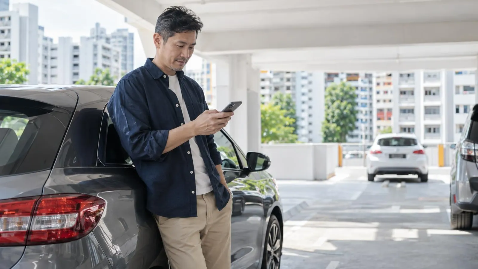 A man leaning on a compact grey car in a Singapore HDB carpark while checking his phone
