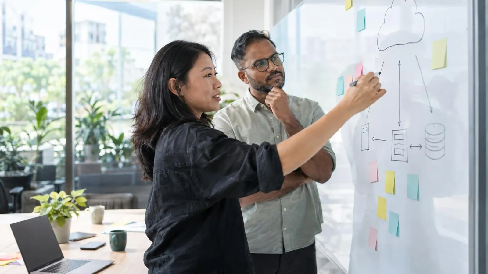 Two IT consultants whiteboarding a cloud migration plan with sticky notes in a bright modern meeting room