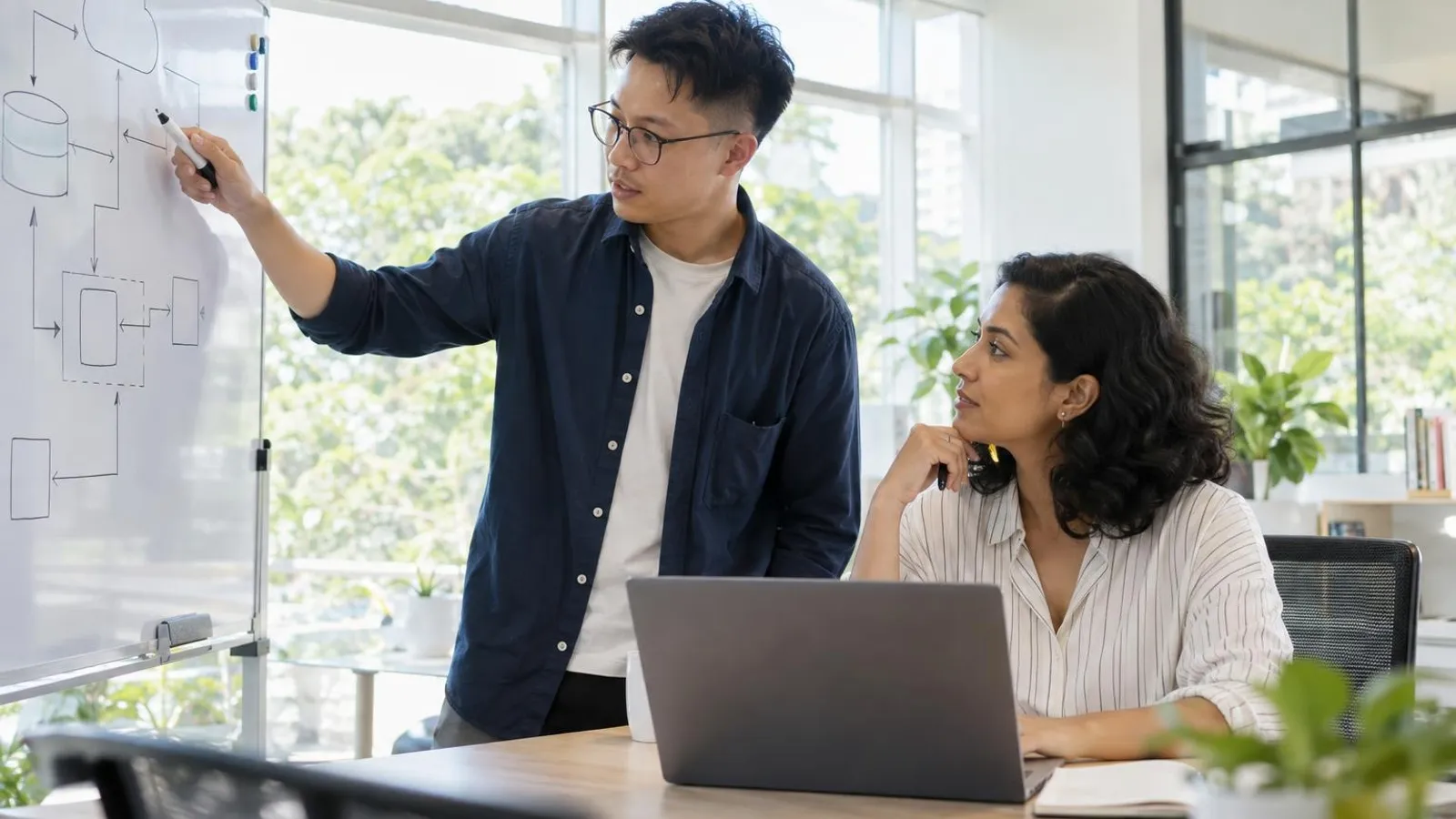 A data engineer and a manager reviewing an abstract data-flow diagram on a whiteboard and laptop in a bright meeting room