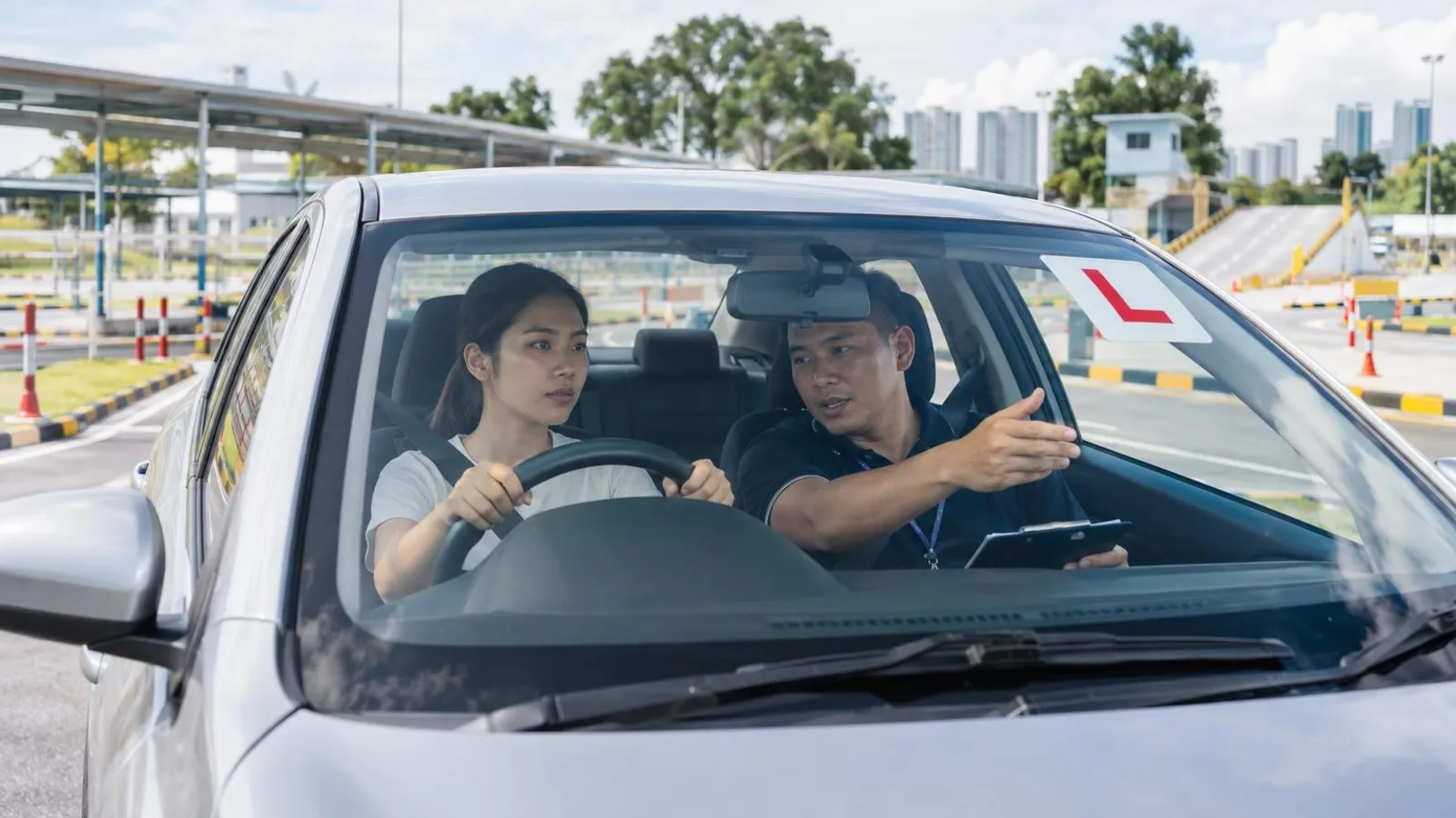 A driving instructor guiding a learner driver in a car at a Singapore driving circuit with an L-plate on the windscreen