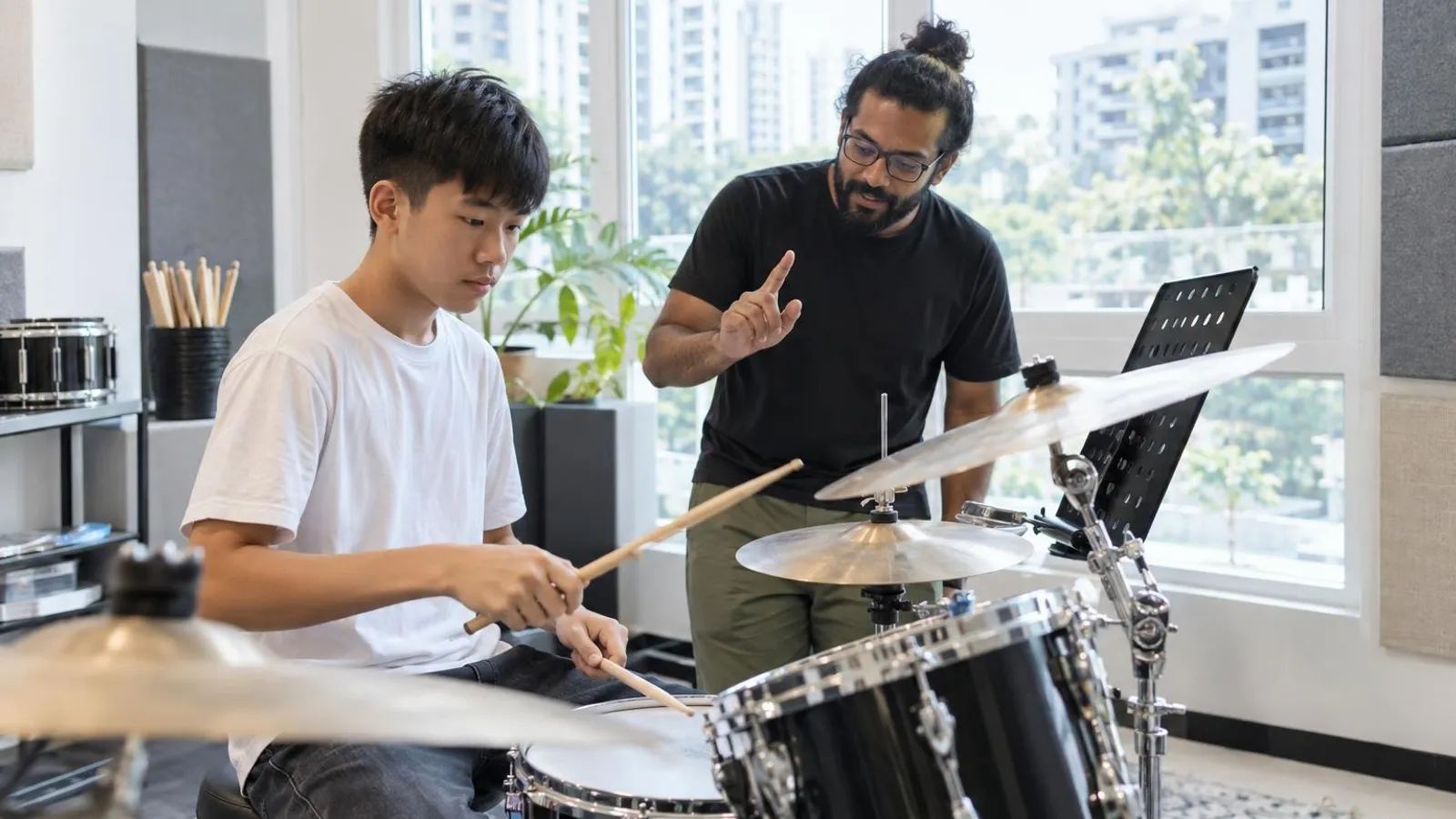 A teenage boy playing a drum kit as his teacher counts the beat in a bright music studio