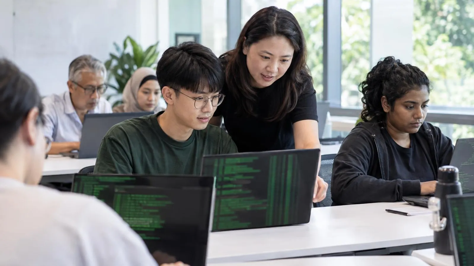 Adult learners at laptops with terminal windows in a bright classroom while an instructor helps
