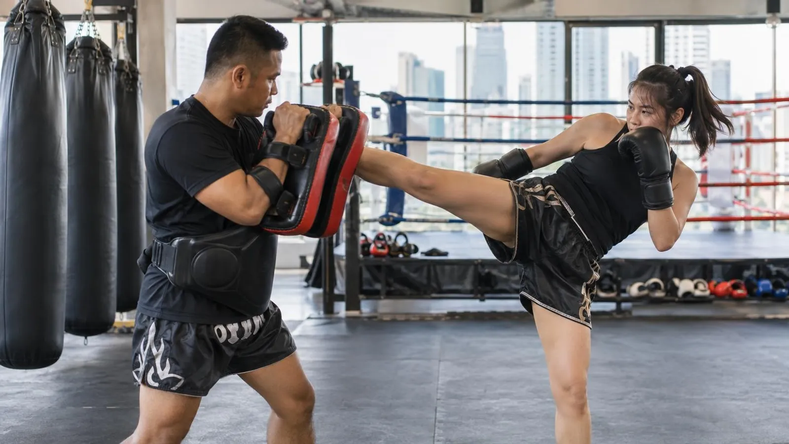A Muay Thai trainer holding pads as a student throws a kick in a bright Singapore gym with a boxing ring