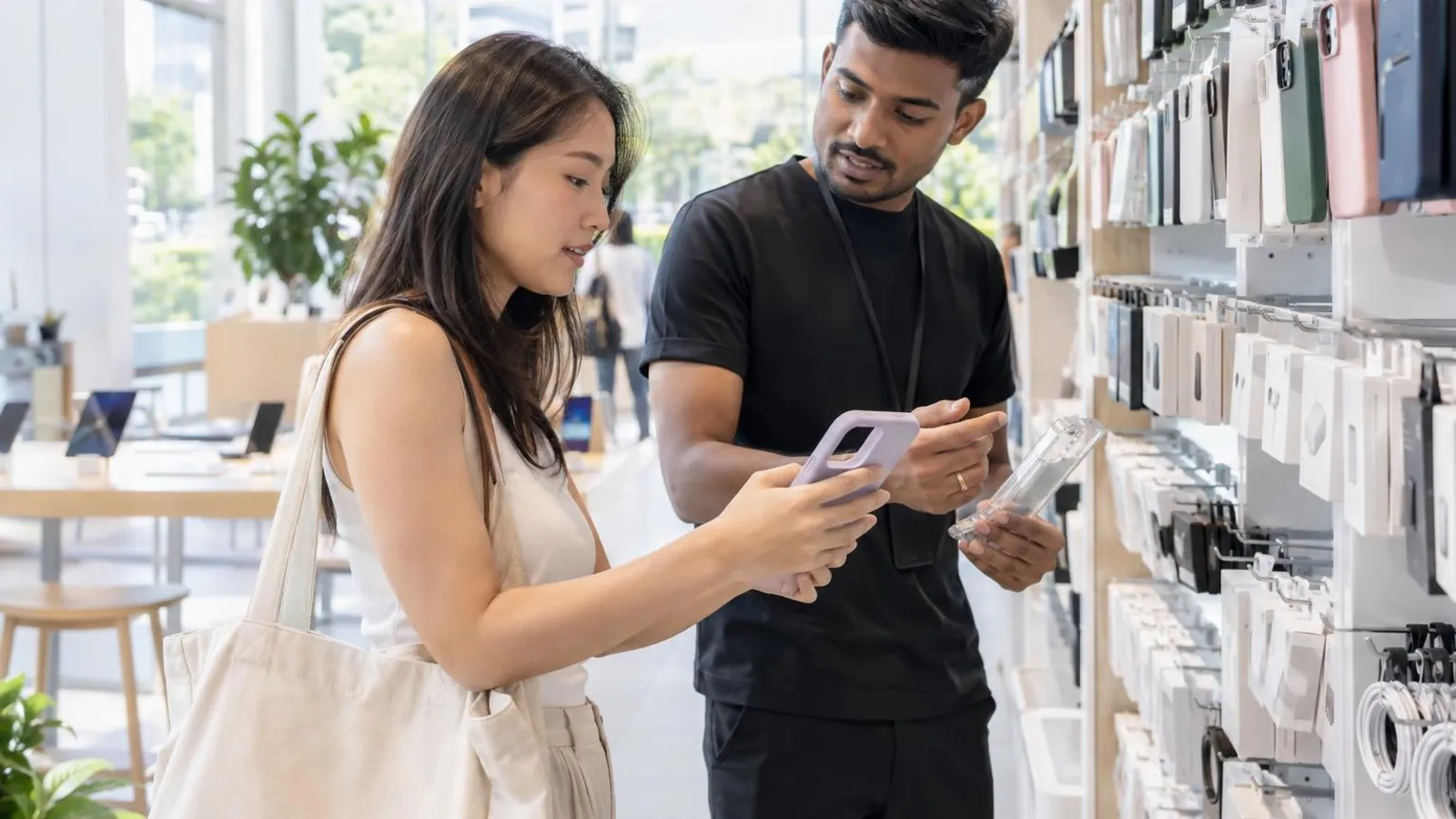 A customer browsing phone cases and chargers on a bright retail wall while a shop assistant helps