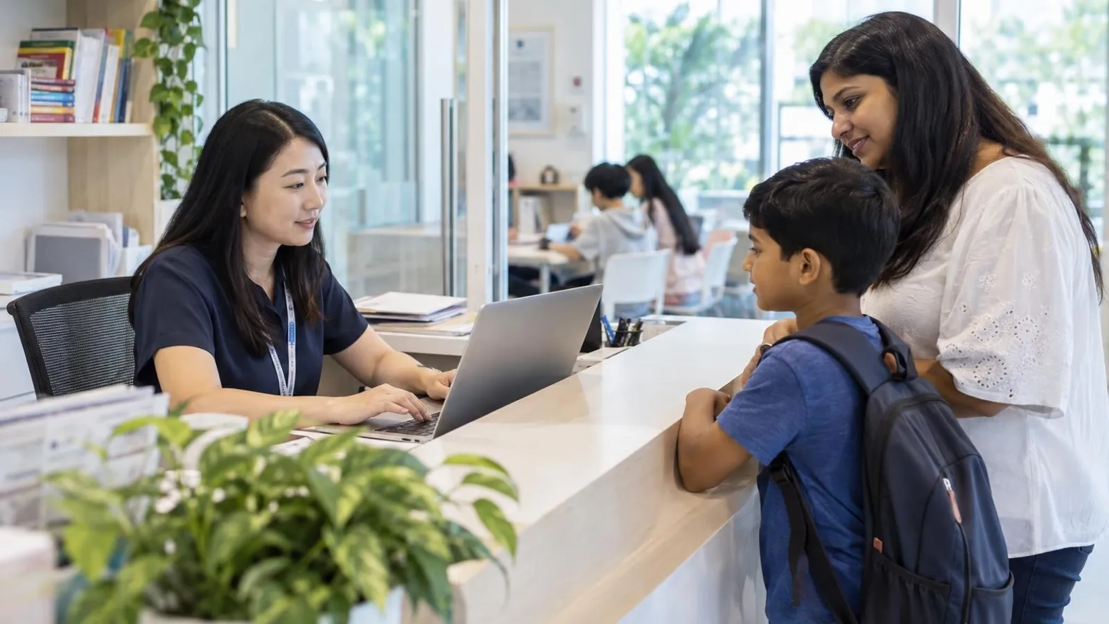 A tuition centre administrator at a bright front desk using a laptop while a parent and child wait