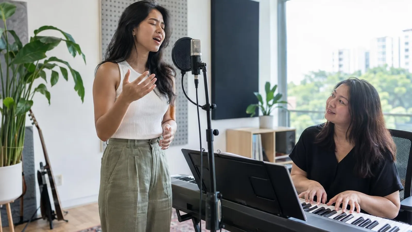 A woman singing beside a vocal coach at a keyboard in a bright studio with a microphone