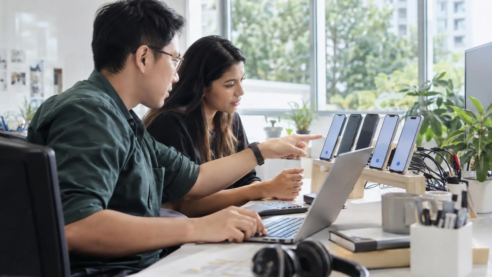 Two software testers checking an app on several phones and a laptop at a bright desk