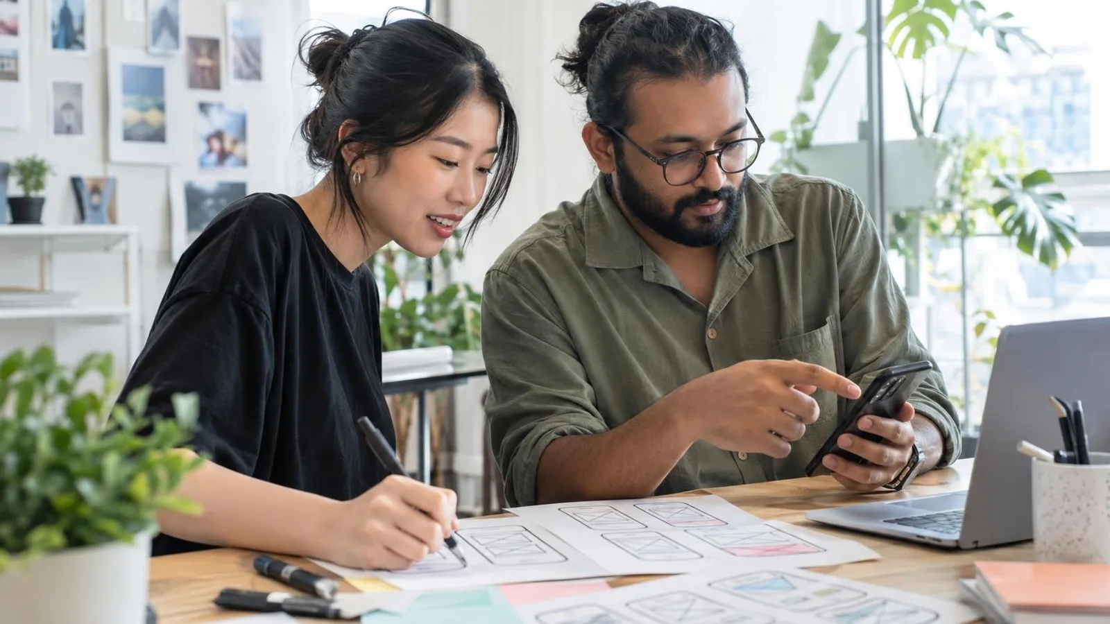 Two designers sketching app screens and testing a clickable prototype on a phone at a bright studio desk